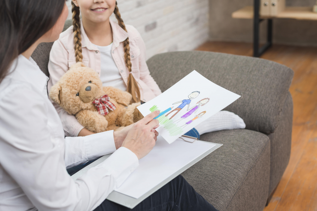 Eine Frau zeigt einem jungen Mädchen, das auf einem Sofa sitzt und einen Teddybär hält, die Zeichnung einer Familie.